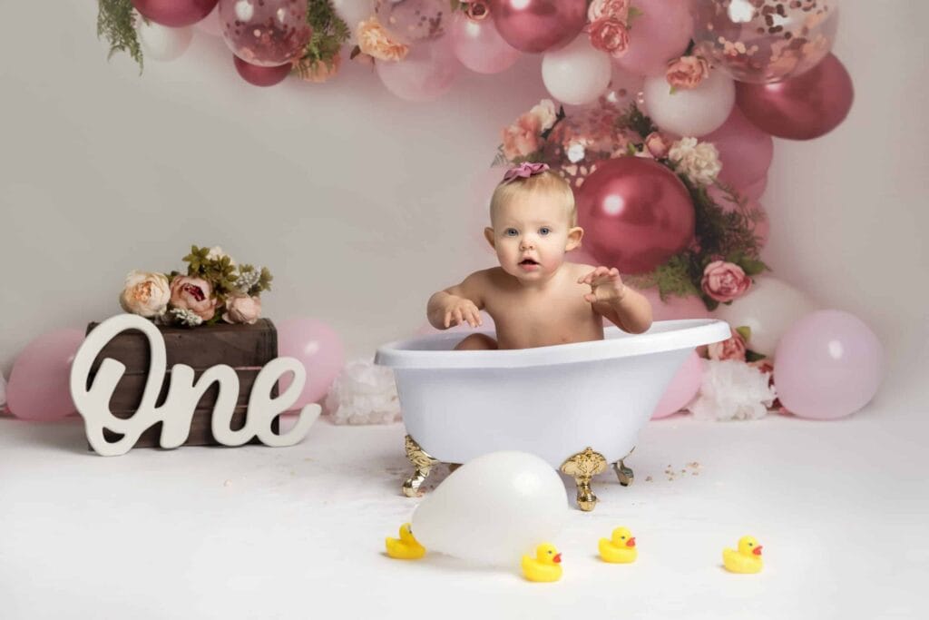 A baby sits in a small white tub surrounded by pink balloons, flowers, rubber ducks, and a sign that reads "One. Drawn by Light Photography