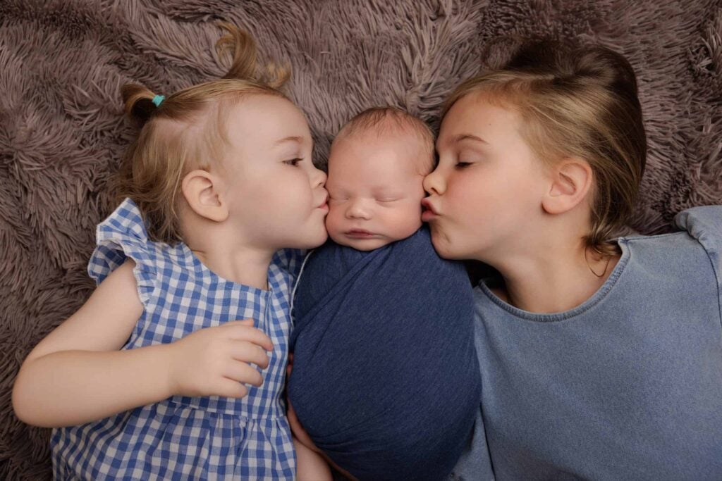 Two young children gently kiss a swaddled newborn baby, all lying on a soft, textured surface. Drawn by Light Photography