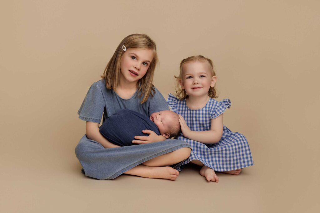 Three young children sit together on a beige background; an older girl holds a sleeping baby whilst a toddler sits beside them, smiling at the camera. Drawn by Light Photography