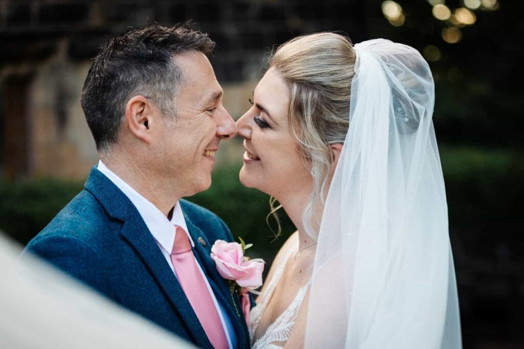 A bride and groom stand close together outdoors, smiling at each other. The bride wears a veil and white dress; the groom wears a blue suit with a pink tie and buttonhole. Drawn by Light Photography
