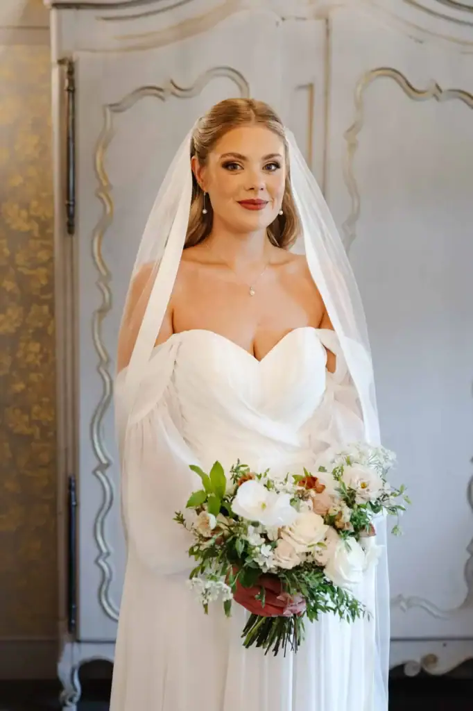 A bride in a white, off-the-shoulder wedding dress holds a bouquet of white and blush flowers, standing indoors in front of a panelled cupboard. Drawn by Light Photography