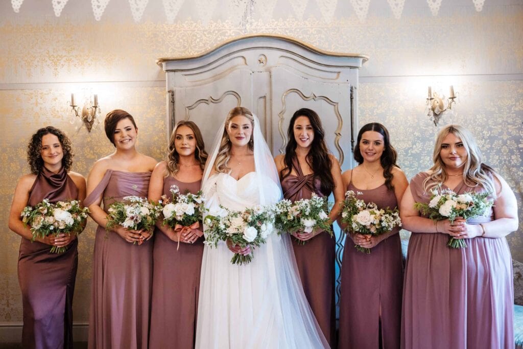 Seven women stand indoors, six in mauve dresses holding bouquets, and one in a white wedding dress and veil, all posing in front of a decorative wardrobe. Drawn by Light Photography