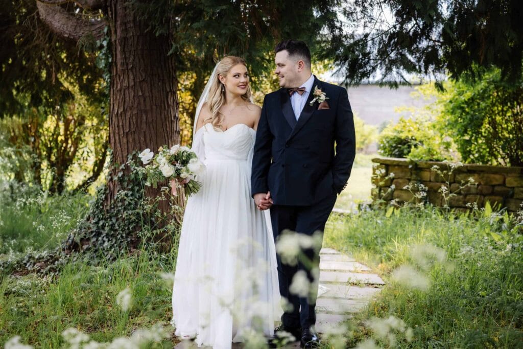 A bride in a white dress and a groom in a black suit walk hand in hand on a garden path, surrounded by greenery and trees. Drawn by Light Photography