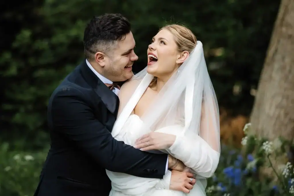 A bride in a white dress and veil smiles and laughs as she is embraced by a groom in a black suit, standing outdoors among greenery and flowers. Drawn by Light Photography
