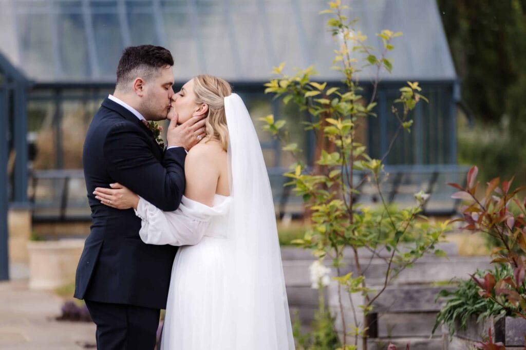A bride and groom in formal wedding attire kiss outdoors in front of a glass conservatory, surrounded by greenery. Drawn by Light Photography