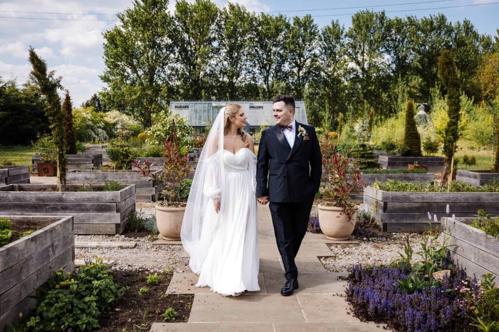 A bride and groom walk hand in hand down a garden path surrounded by greenery and raised wooden planters on a sunny day. Drawn by Light Photography