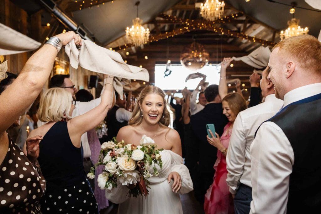 A bride holding a bouquet walks through a crowd of guests waving serviettes in a decorated indoor venue with chandeliers and fairy lights. Drawn by Light Photography