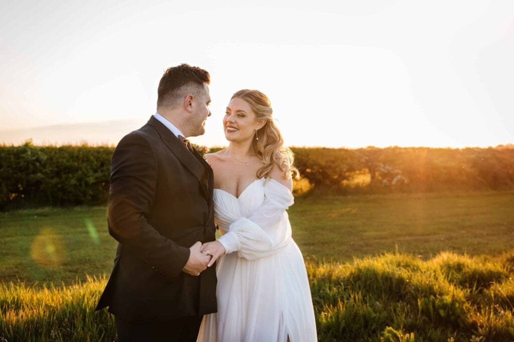 A bride and groom stand outdoors at sunset, holding hands and looking at each other, with green grass and a hedge in the background. Drawn by Light Photography