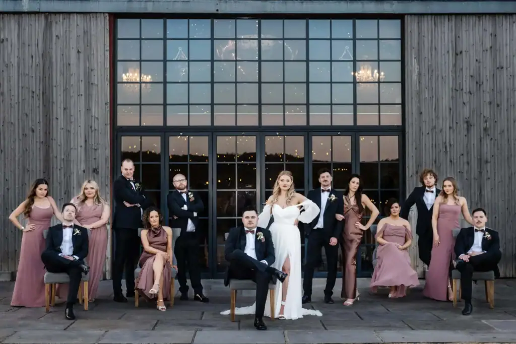 A wedding party poses in formal attire in front of a large glass and wood building; the bride and groom are seated at the centre, surrounded by bridesmaids and ushers. Drawn by Light Photography