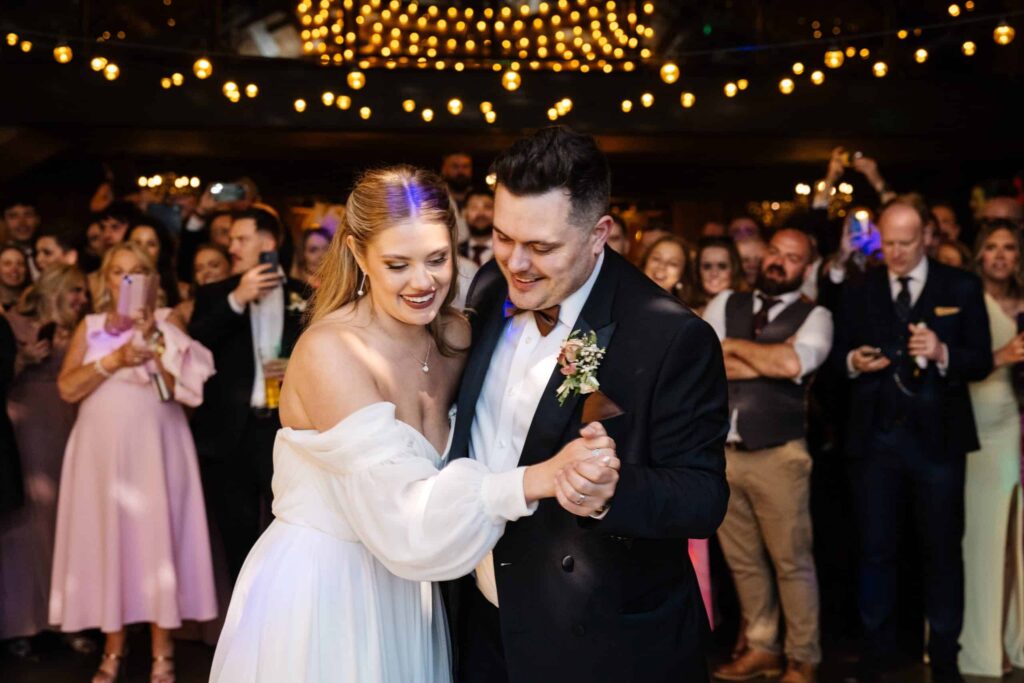 A bride and groom dance together at their wedding reception, surrounded by guests under warm fairy lights. Drawn by Light Photography
