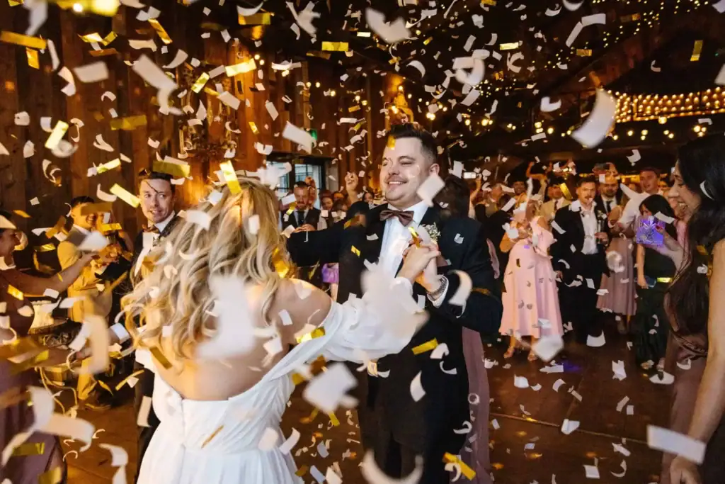 A bride and groom dance surrounded by guests and falling confetti in a warmly lit indoor venue. Drawn by Light Photography