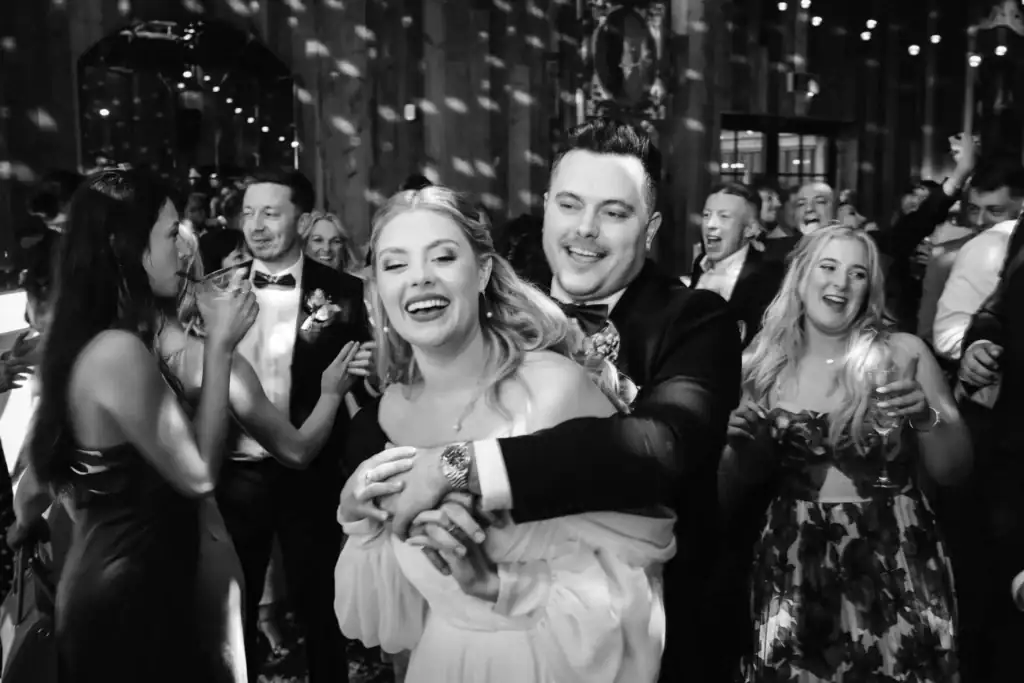 A couple in formal attire embraces and smiles on a crowded dance floor at a lively indoor event, surrounded by other guests in evening dress. Drawn by Light Photography