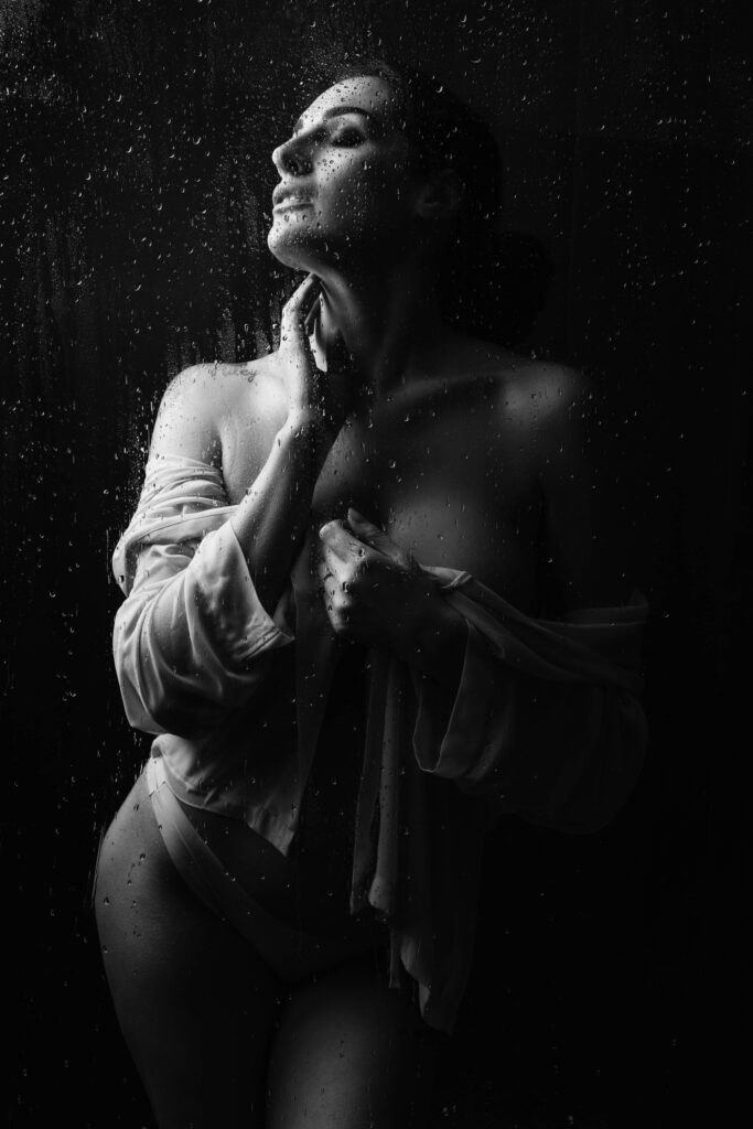 Black and white photo of a woman standing behind wet glass, wearing an open shirt and underwear, with one hand on her neck and looking upwards. Drawn by Light Photography