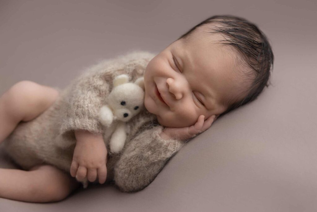 A newborn baby in a cosy outfit smiles whilst lying on its side, resting its head on one hand and holding a small white teddy bear. Drawn by Light Photography