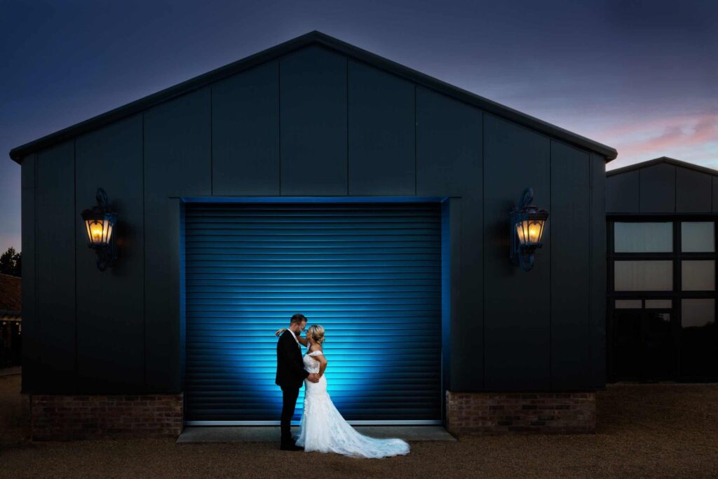 A bride and groom embrace in front of a large industrial building with a blue-lit garage door at dusk. Drawn by Light Photography