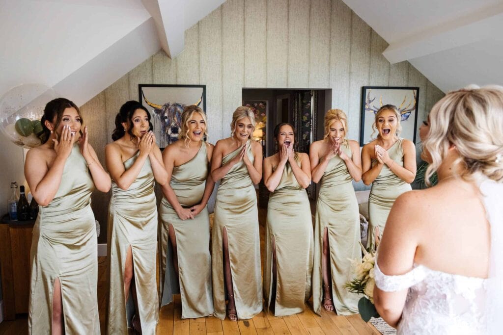Six bridesmaids in matching light green dresses react with excitement as the bride, seen from behind, shows them her wedding dress in a well-lit room. Drawn by Light Photography