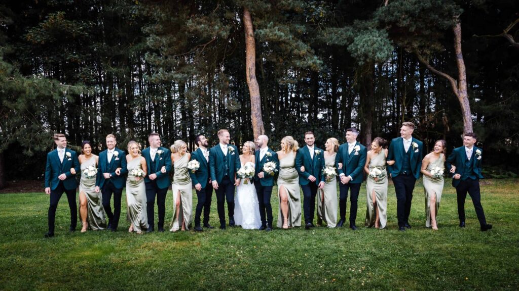 A wedding party in formal attire stands in a queue outdoors, with bridesmaids in sage dresses and groomsmen in dark suits, smiling and linking arms. Trees fill the background. Drawn by Light Photography