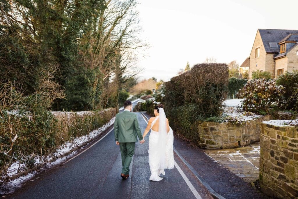 A bride and groom walk hand in hand down a narrow road, bordered by hedges and stone walls, on a winter’s day. Drawn by Light Photography