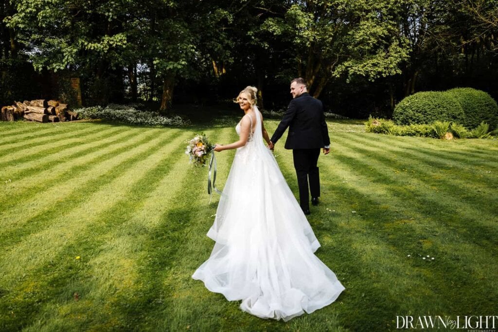 A bride and groom walk hand in hand across a striped green lawn, with trees and shrubs in the background. Drawn by Light Photography