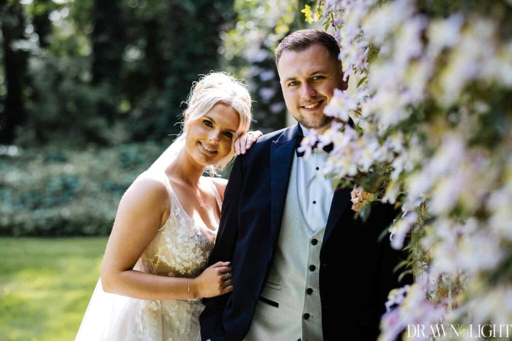 A bride in a white dress and veil stands beside a groom in a dark suit and bow tie, both smiling outdoors next to a wall covered in flowers. Drawn by Light Photography