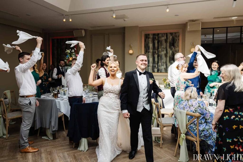 Bride and groom enter a reception hall smiling and holding hands as guests stand and wave serviettes in celebration. Drawn by Light Photography