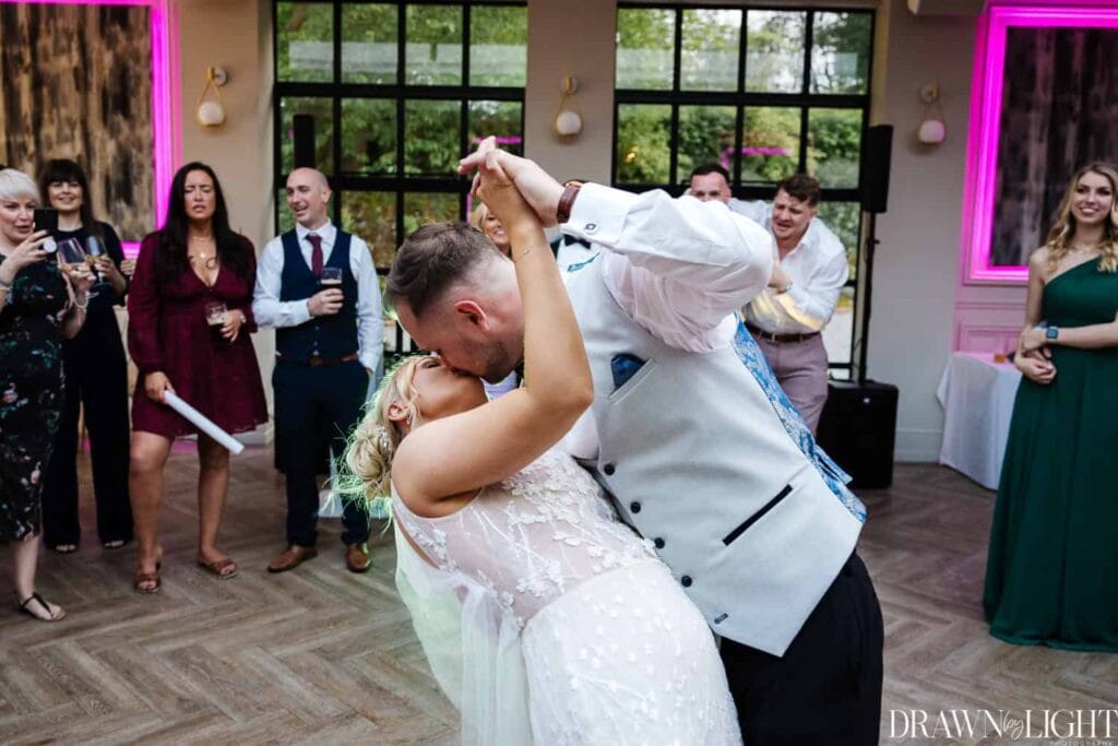 A newlywed couple share a dip and kiss on the dance floor at their wedding reception, surrounded by smiling guests. Drawn by Light Photography