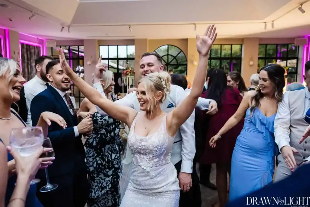 A bride in a white dress dances with guests at a wedding reception, smiling with her arms raised. People around her are laughing and enjoying the celebration. Drawn by Light Photography