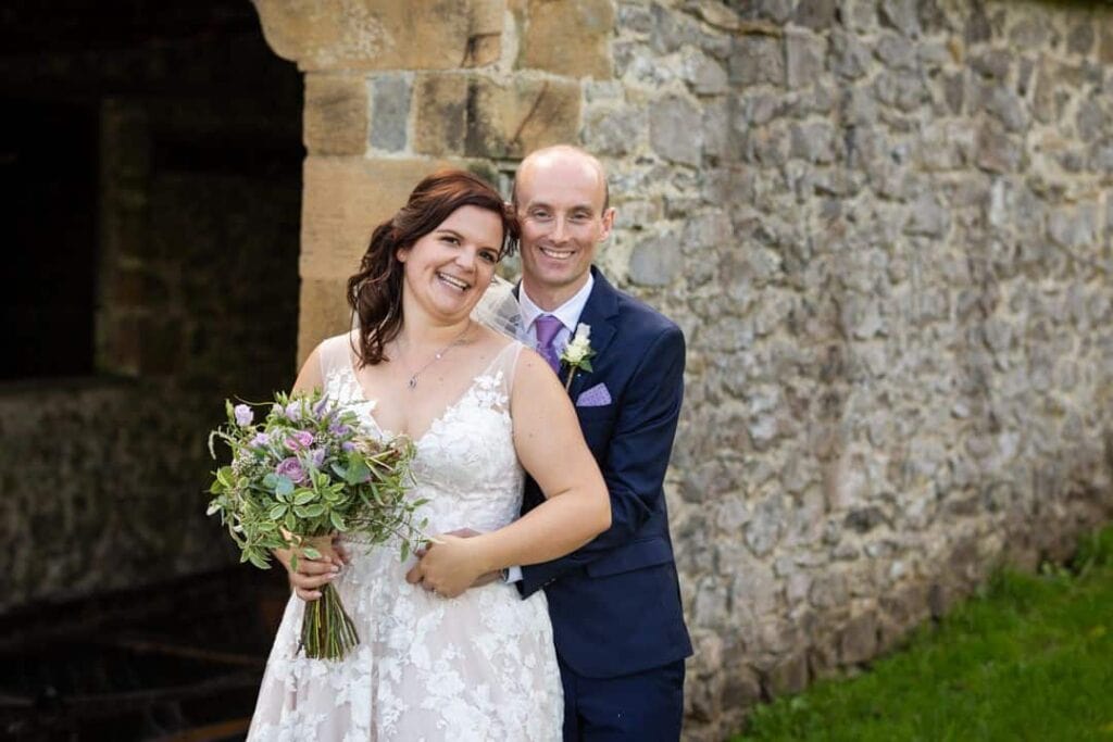 A bride in a white dress holds a bouquet whilst standing in front of a stone wall with a groom in a dark suit standing closely behind her, both smiling at the camera. Drawn by Light Photography