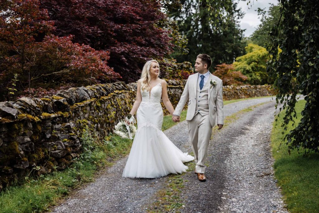 A bride and groom walk hand-in-hand down a gravel path lined with stone walls and greenery, both dressed in formal wedding attire. Drawn by Light Photography