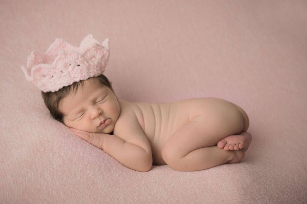 A newborn baby sleeps on a pink blanket, wearing a pink knitted crown, with hands under the cheek and legs curled up. Drawn by Light Photography
