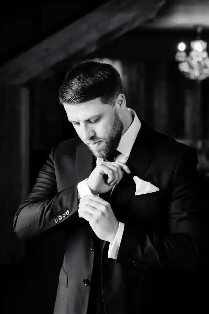 A man in a suit adjusts his cufflink and pocket square, looking down thoughtfully in a dimly lit, elegant setting. Drawn by Light Photography