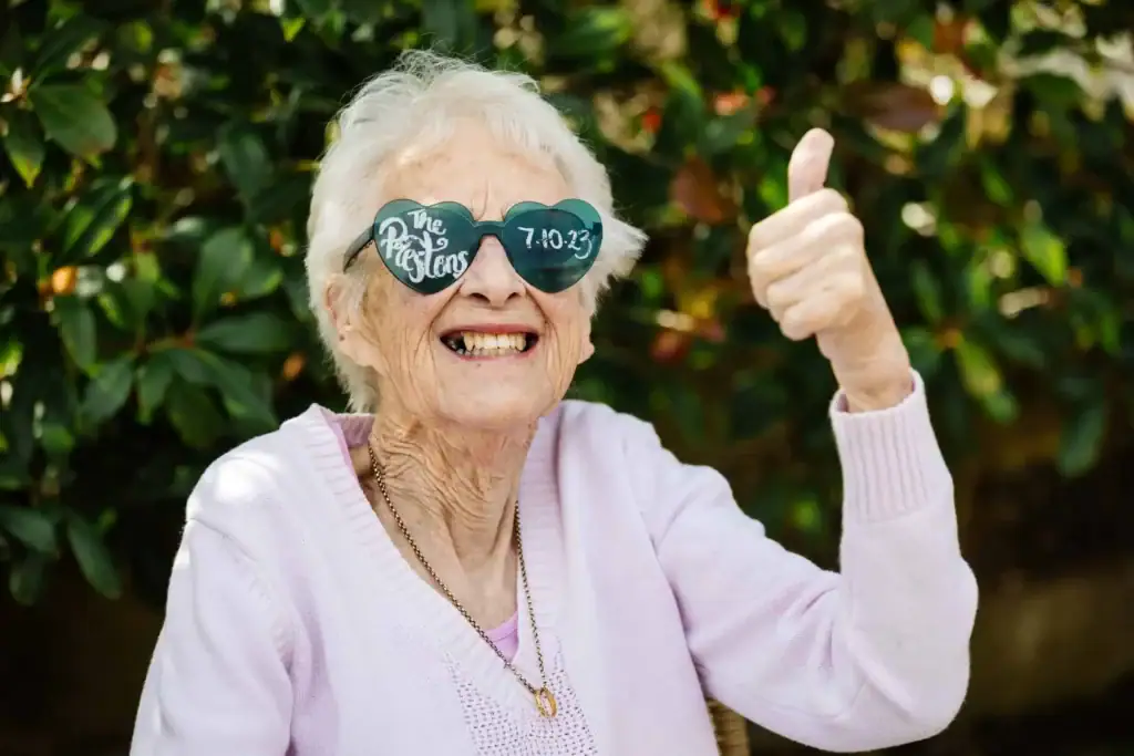 Elderly woman wearing heart-shaped sunglasses with text, smiling and giving a thumbs-up, outdoors with greenery in the background. Drawn by Light Photography