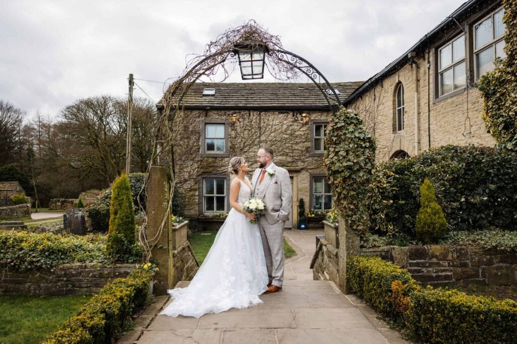 A bride and groom stand facing each other on a stone path outside a rustic building, surrounded by greenery and overcast skies. Drawn by Light Photography