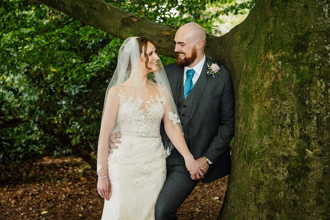 A bride and groom in wedding attire stand together holding hands by a large tree outdoors, smiling at one another. Drawn by Light Photography