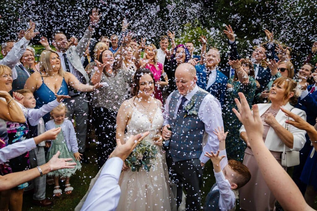 A bride and groom walk outdoors surrounded by guests throwing confetti, with people cheering and celebrating their wedding. Drawn by Light Photography
