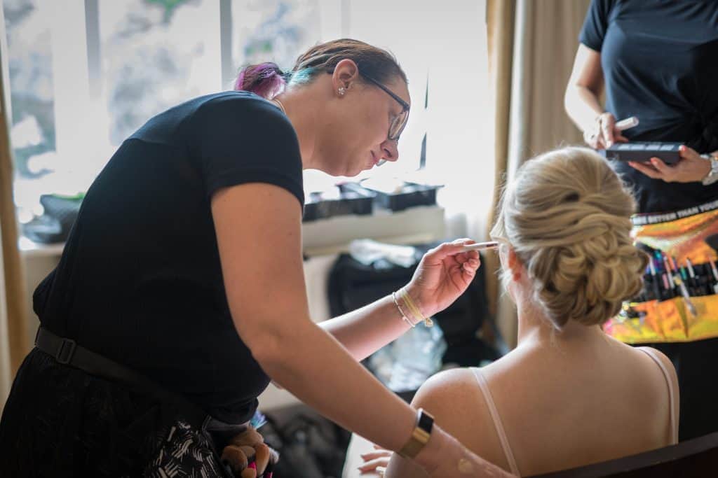 A makeup artist expertly applies bridal make up to a seated woman with blonde hair in an elegant updo, while another person stands nearby holding supplies. Image by Lisa Kershaw - Drawn By Light Photography - West Yorkshire - UK