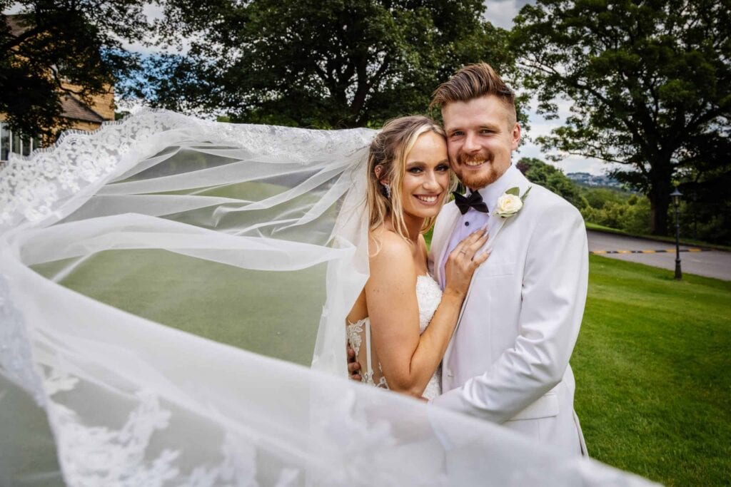 Bride and groom smiling and embracing outdoors, with the bride's veil flowing in the foreground and green trees in the background. Drawn by Light Photography