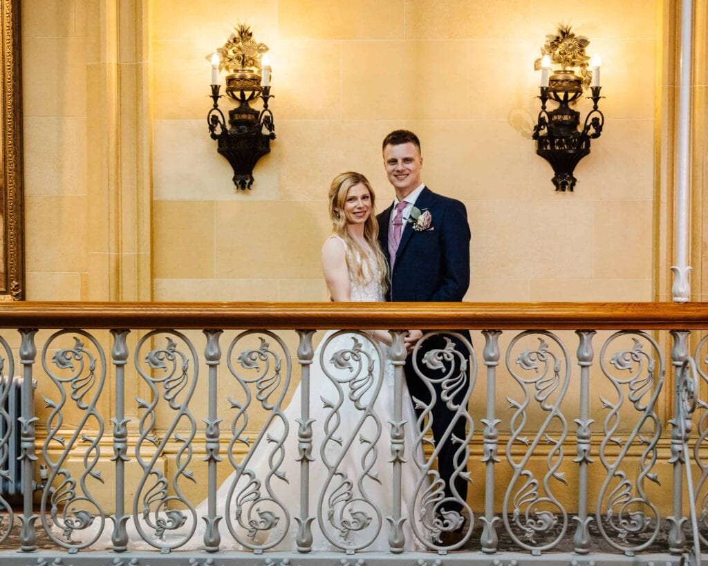 A bride and groom stand side by side in formal attire, smiling, behind an ornate railing with two wall sconces lit behind them. Drawn by Light Photography