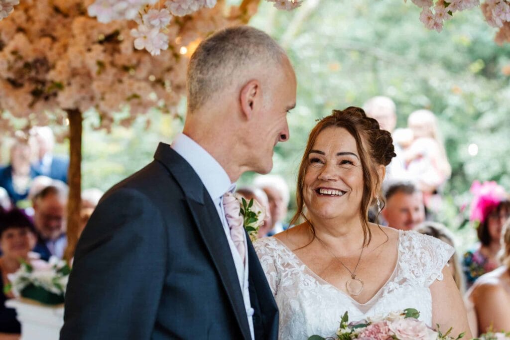 A bride and groom stand together at their wedding ceremony, smiling at each other, with guests and floral decorations in the background. Drawn by Light Photography