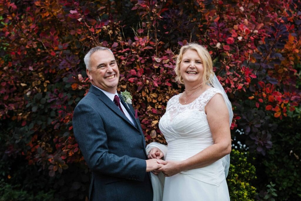 A bride and groom stand outdoors, holding hands and smiling, with colourful autumn foliage in the background. Drawn by Light Photography