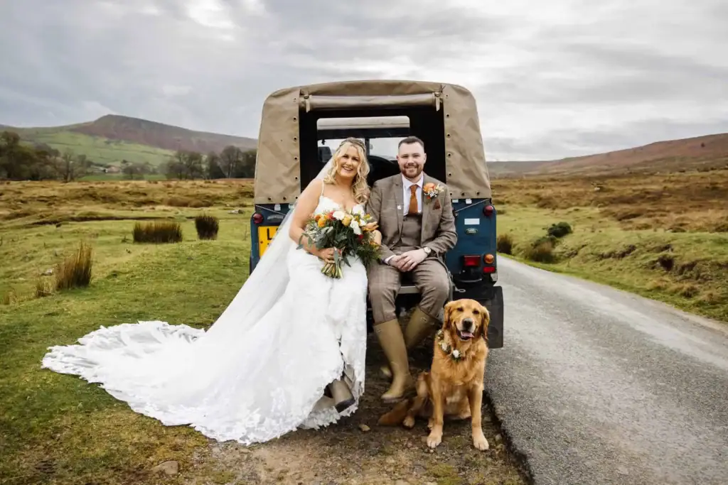 A bride and groom sit on the back of a jeep, holding a bouquet, with a golden retriever beside them on a country road, green hills in the background. Drawn by Light Photography