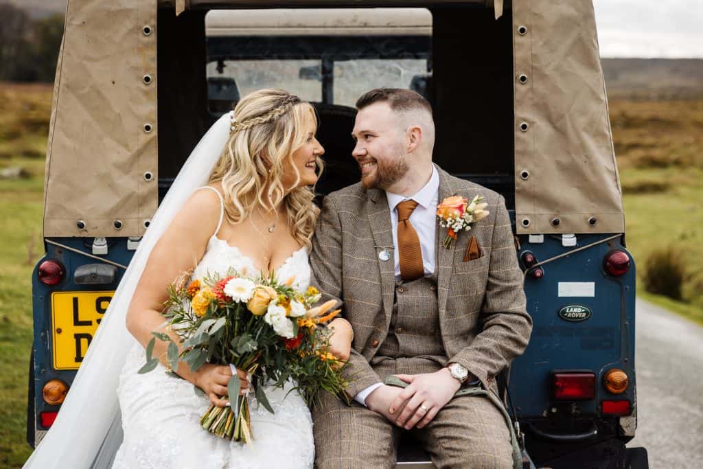 Smiling newlyweds sitting on Land Rover tailgate