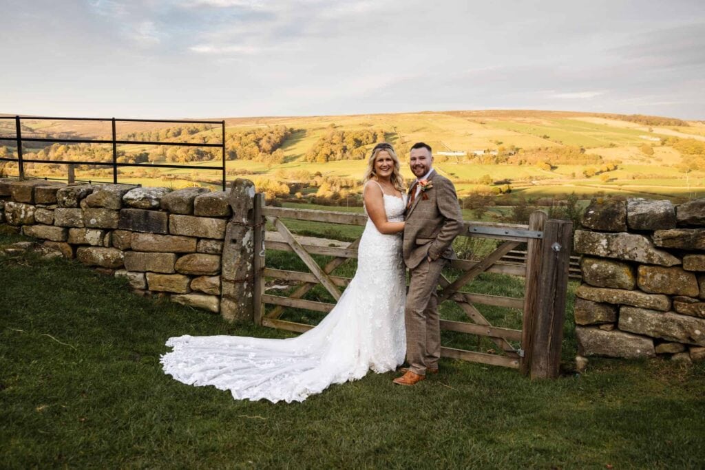 Bride and groom pose together by a wooden gate and stone wall in a rural landscape, with green fields and hills in the background. Drawn by Light Photography