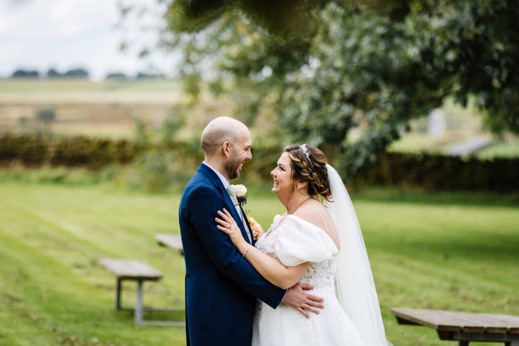 A bride and groom stand outside on grass, embracing and smiling at each other, with benches and trees in the background. Drawn by Light Photography