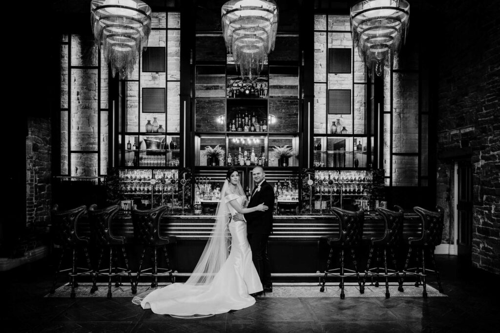 A bride and groom stand together in front of a bar inside an elegant venue, with high-backed chairs and tall windows, in a black and white photograph. Drawn by Light Photography
