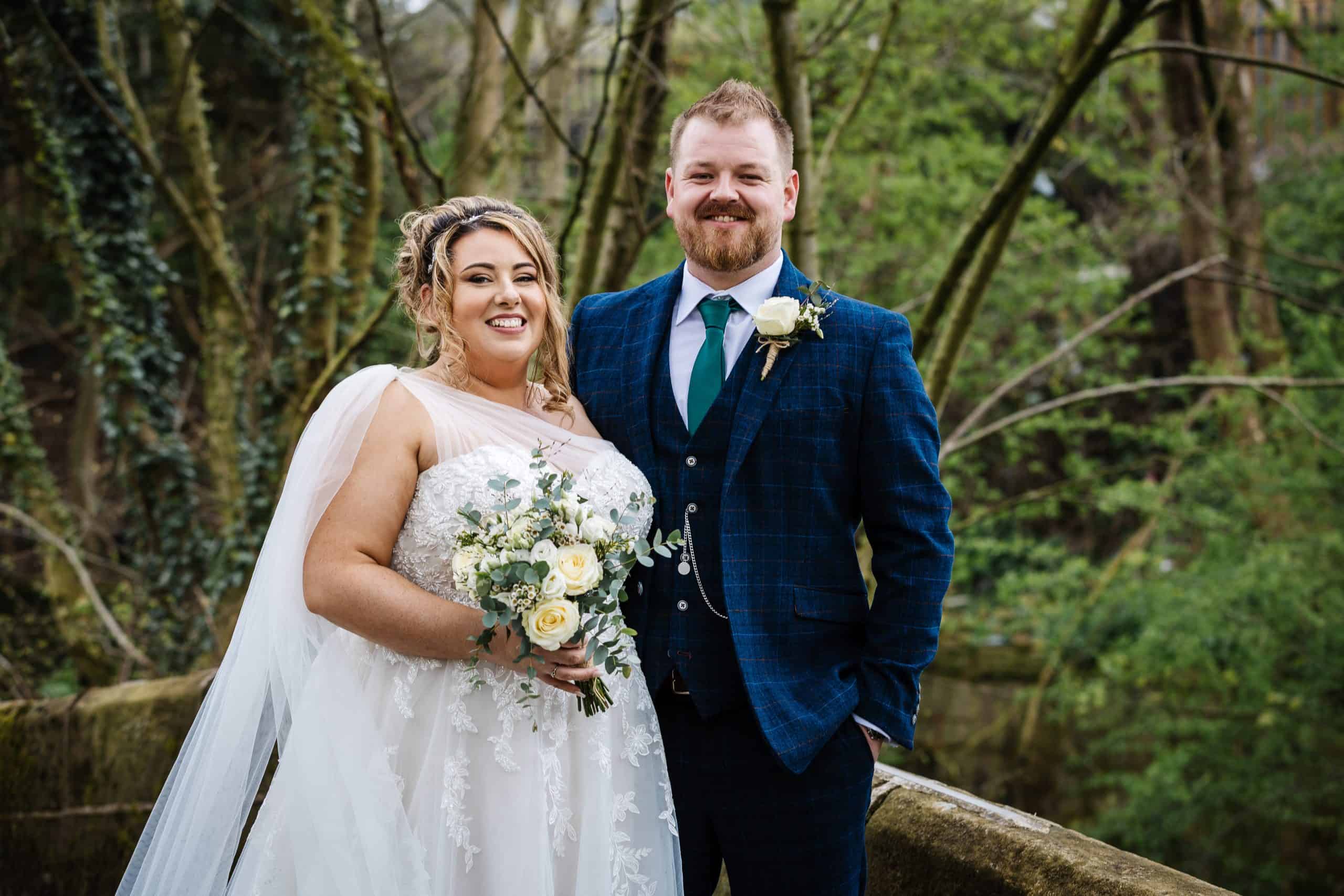 A bride in a white dress and a groom in a blue suit stand outdoors, smiling and holding a bunch of white and yellow flowers. Trees are visible in the background. Drawn by Light Photography