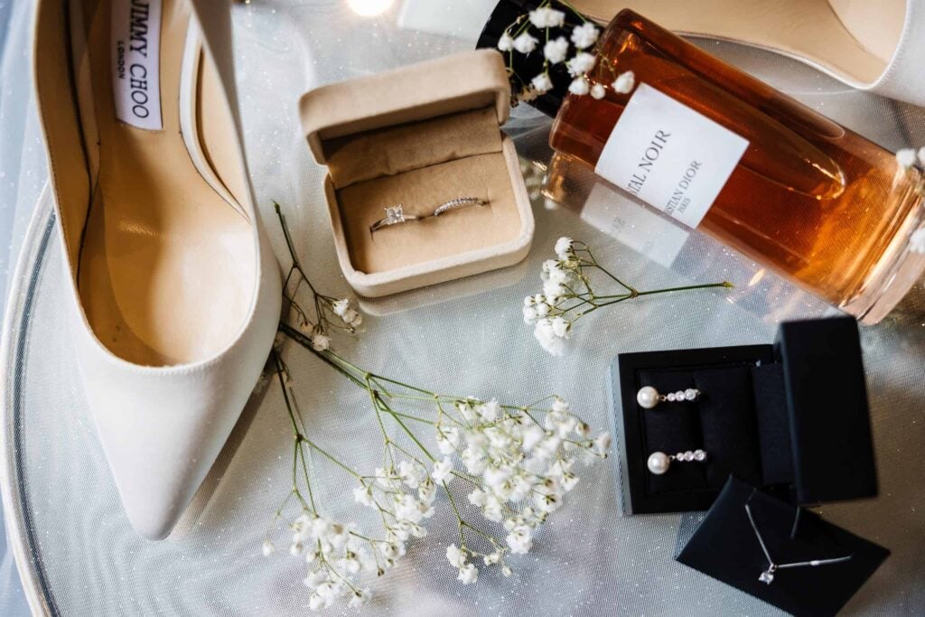 White high-heeled shoe, engagement ring in box, perfume bottle, pearl earrings, necklace, and baby's breath flowers arranged on a reflective surface. Drawn by Light Photography