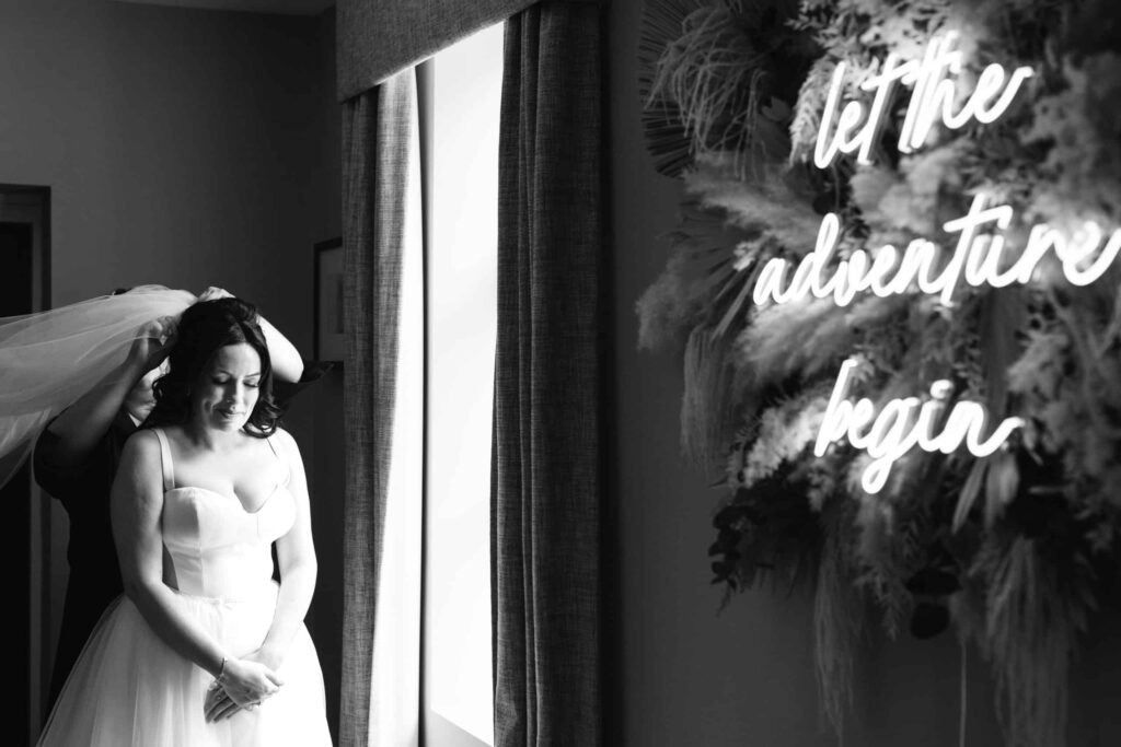 A bride stands by a window as someone adjusts her veil; a neon sign beside her reads "let the adventure begin. Drawn by Light Photography