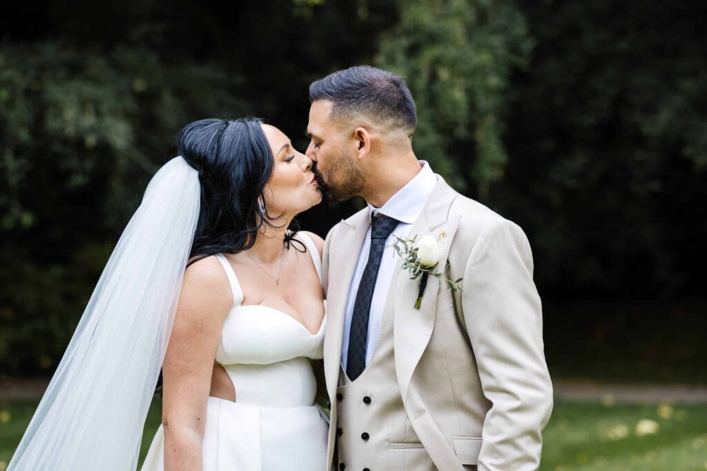 A bride and groom dressed in wedding attire share a kiss outdoors, with greenery in the background. Drawn by Light Photography