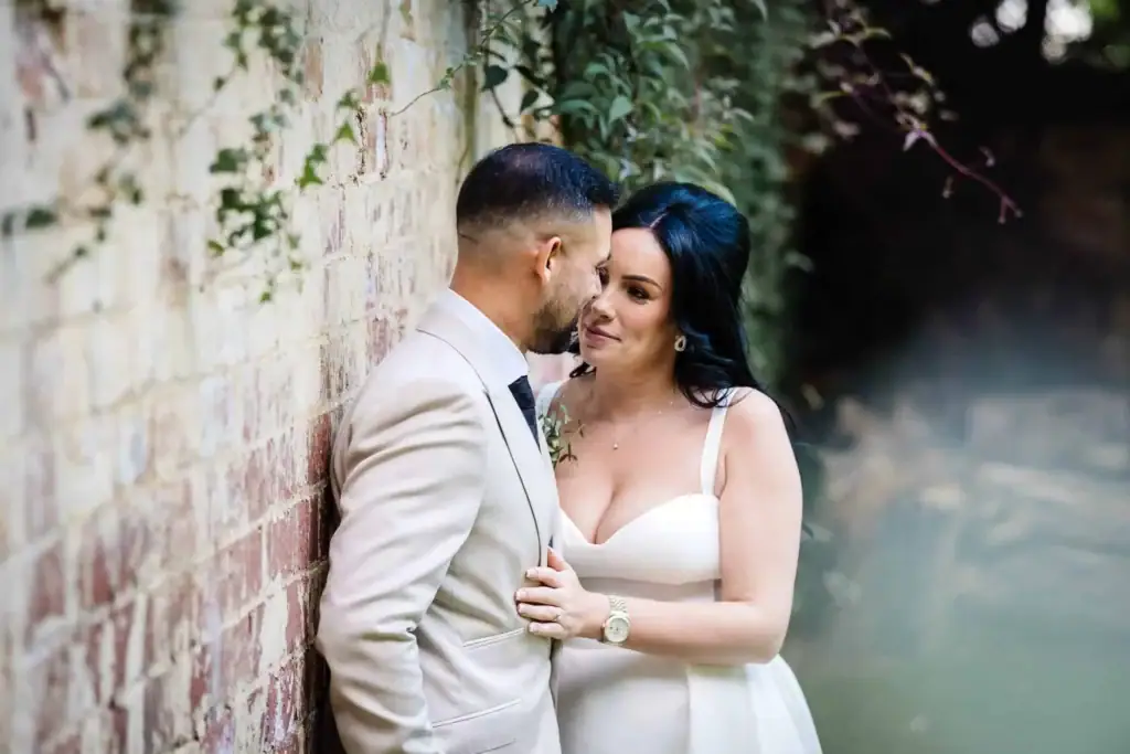 A bride and groom stand closely by a brick wall, facing each other and sharing an intimate moment outdoors. Drawn by Light Photography